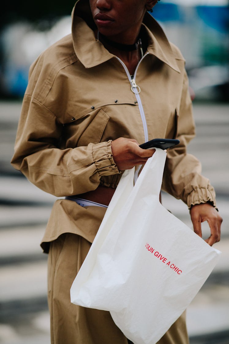 A woman carrying a white plastic bag while wearing a brown jacket and brown baggy pants 