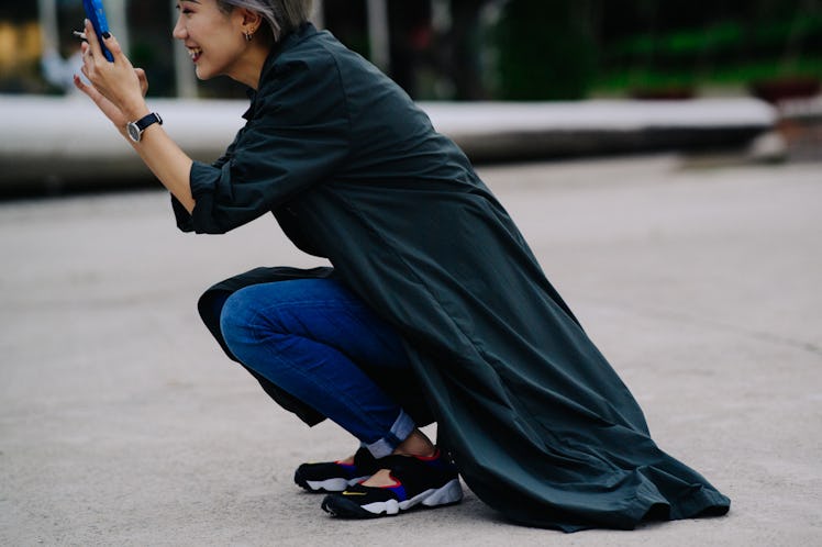 A woman taking a photo on her phone while crouching and wearing a black cardigan and blue pants