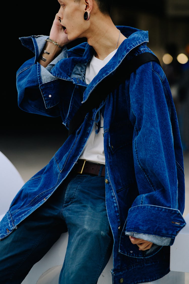 A man talking on his phone while wearing a blue jacket, blue pants, a white shirt, and black earring...