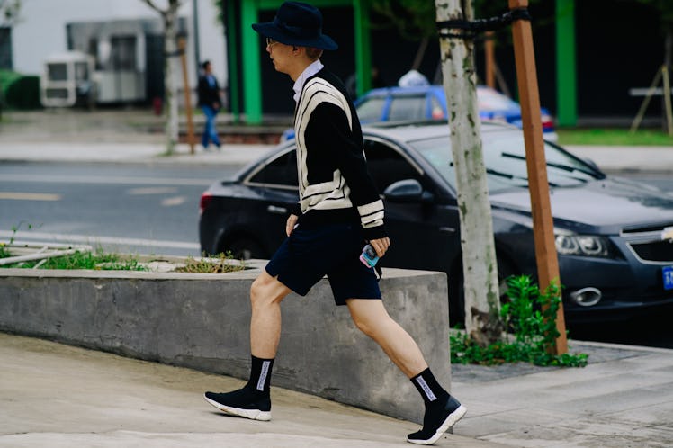 A man walking while wearing black shorts, a black and white shirt, and a black bucket hat