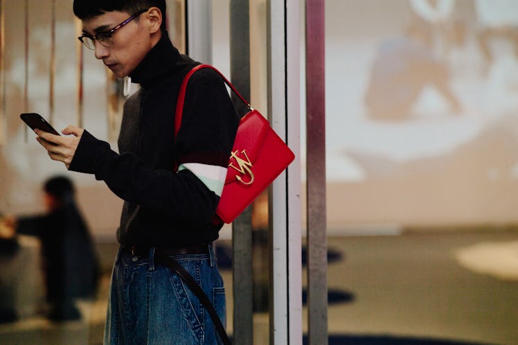 A man using a phone while standing and wearing a black turtleneck and a red handbag