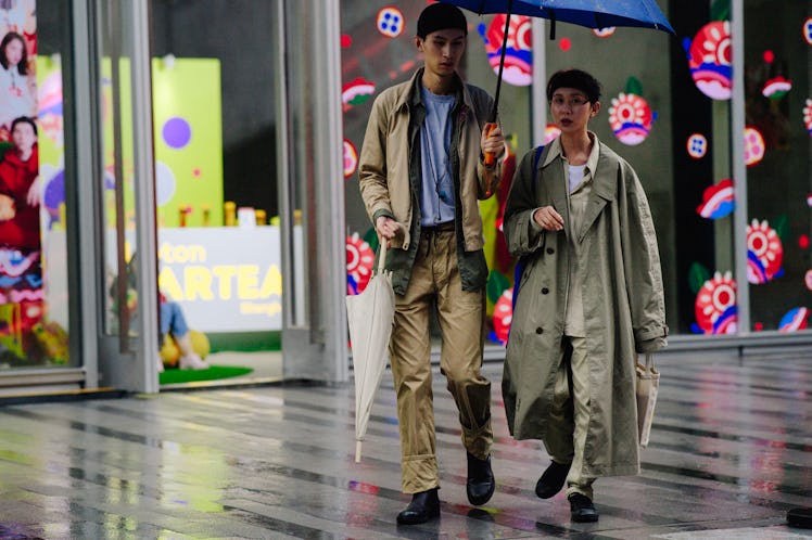 A couple walking under an umbrella, a man is wearing a brown jacket and brown pants while the lady i...