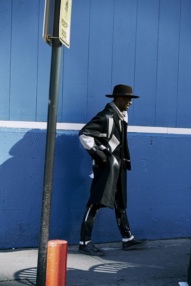 A man walking down a street in a black combination of a coat, hat, shoes, and pants