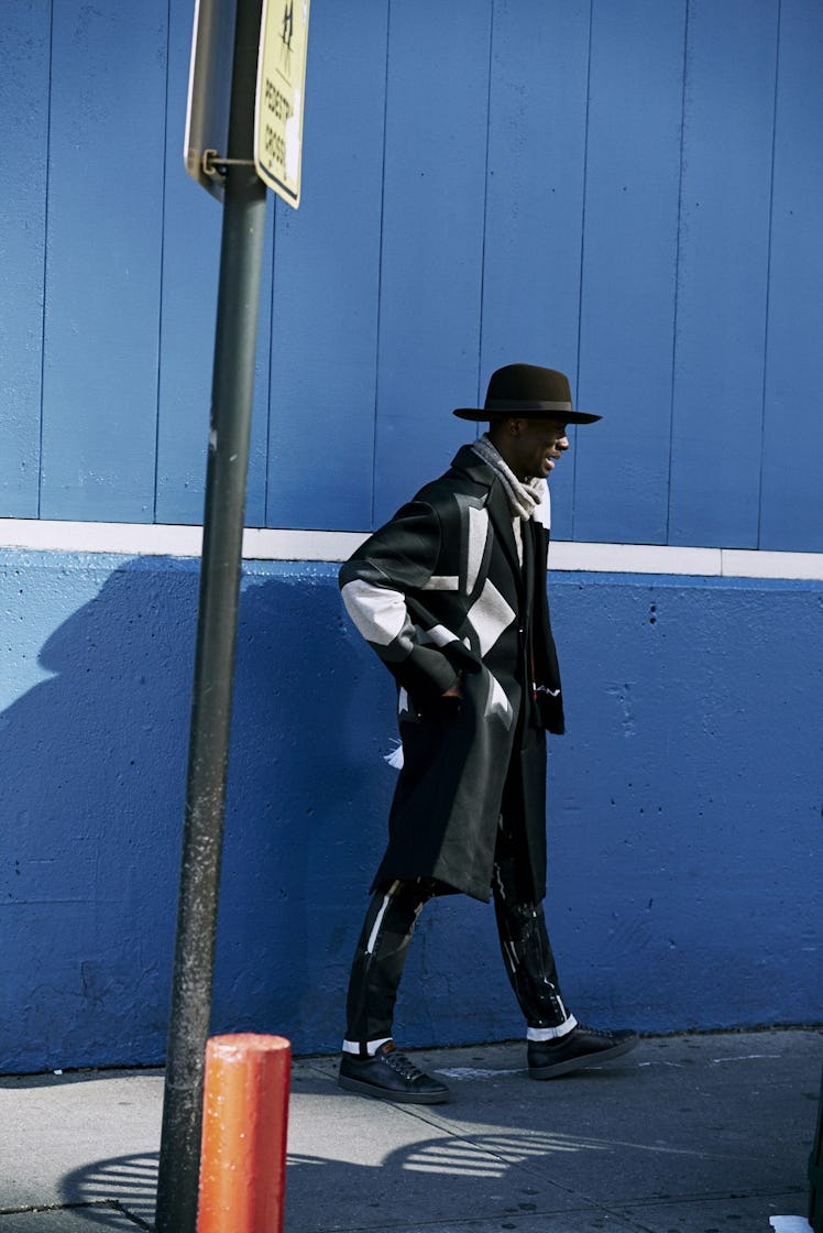 A man walking down a street in a black combination of a coat, hat, shoes, and pants