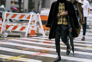 A model crossing a street while wearing a black coat, yellow plaid shirt, and black pants