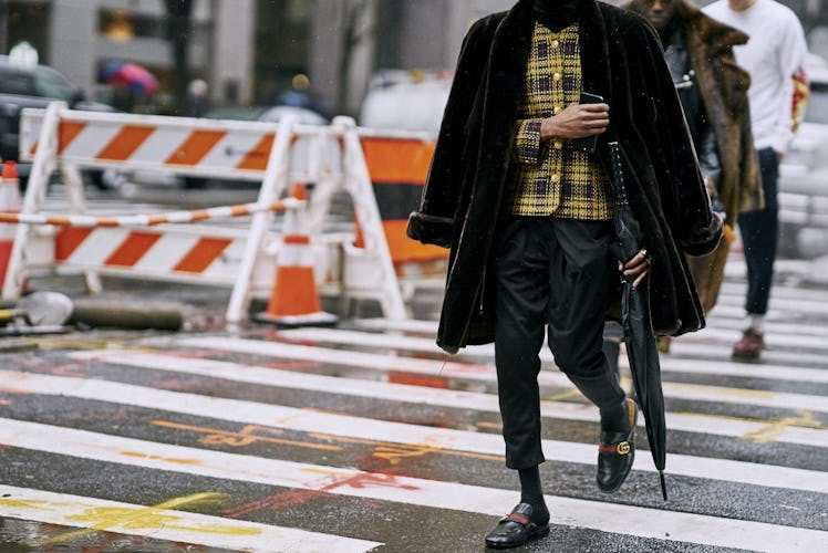 A model crossing a street while wearing a black coat, yellow plaid shirt, and black pants