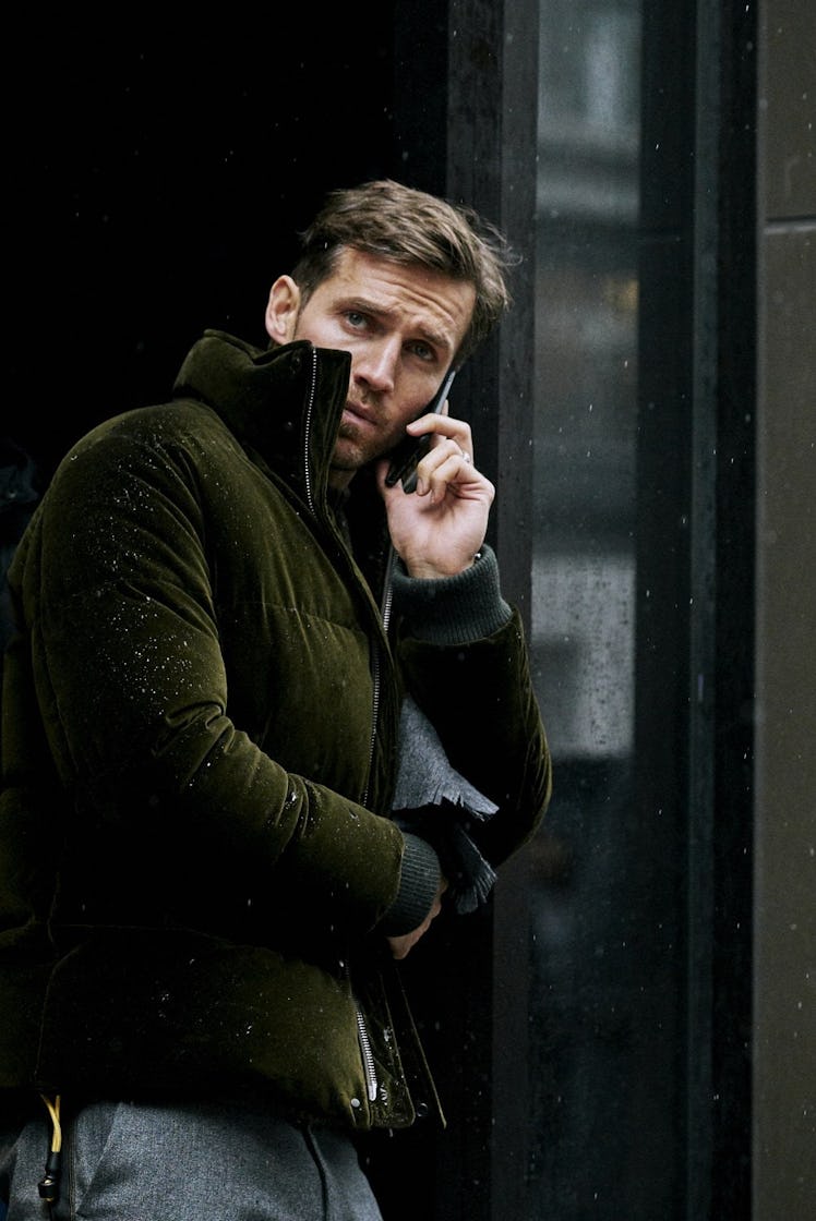 A man walking down a street and talking on his mobile phone while wearing a green jacket