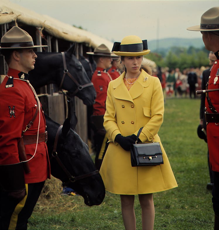 Princess Anne wearing a retro yellow coat next to some guards and horses