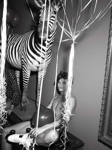 A nude model covering his private body parts with a balloon sitting next to a zebra statue during a ...