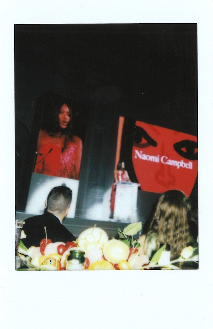 A man and a woman sitting at a table with fruits on it and Naomi Campbell giving a speech on the sta...