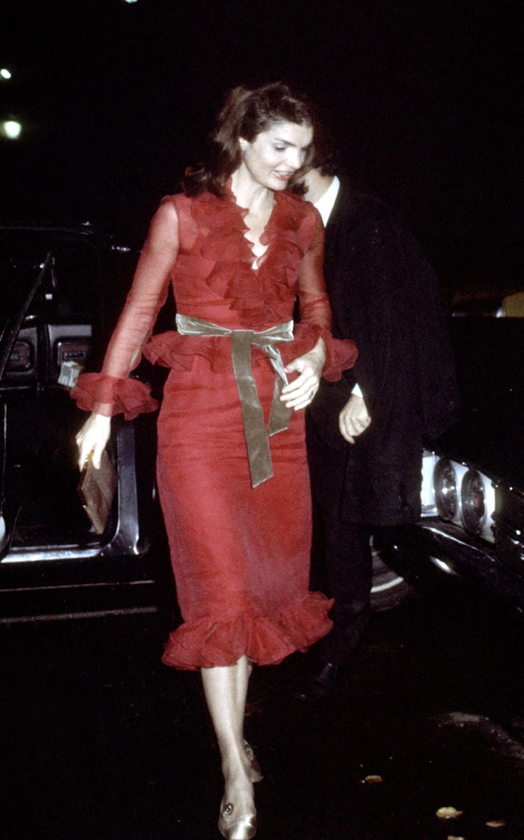 Jackie O wearing a frilly red gown with a thick golden belt an a brown leather tote, sporting an up-...