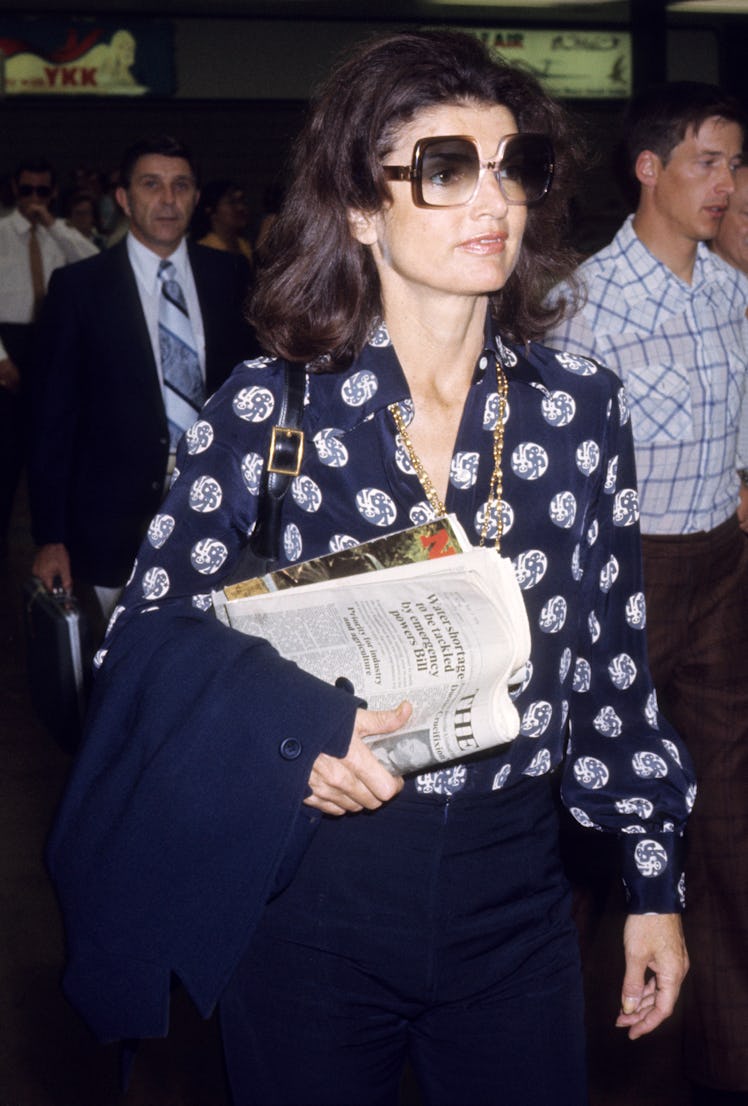 Jackie O at Heathrow Airport in a loose-fitting blouse, high-wasted pants and oversized sunglasses.