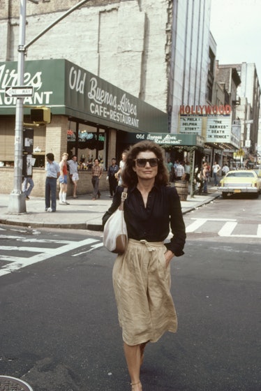 Jackie O in front of a cinema wearing a black blouse, tucked into a beige skirt and oversized sungla...