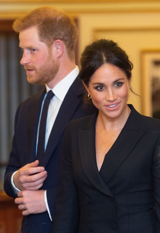 Meghan Markle wearing a black tuxedo dress while standing next to Prince Harry and looking to the si...