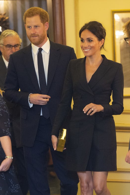 Meghan Markle wearing a black tuxedo dress while smiling and standing next to Prince Harry at a gala...