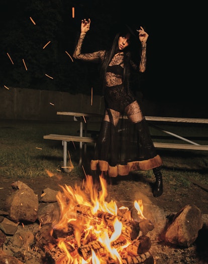 A woman in a black dress posing in front of a bonfire like a witch.