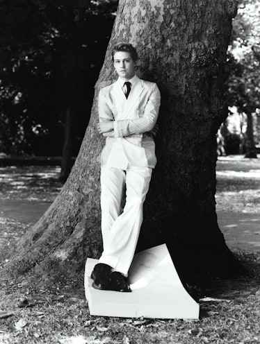 Joe Alwyn posing in a white formal suit while leaned against a tree