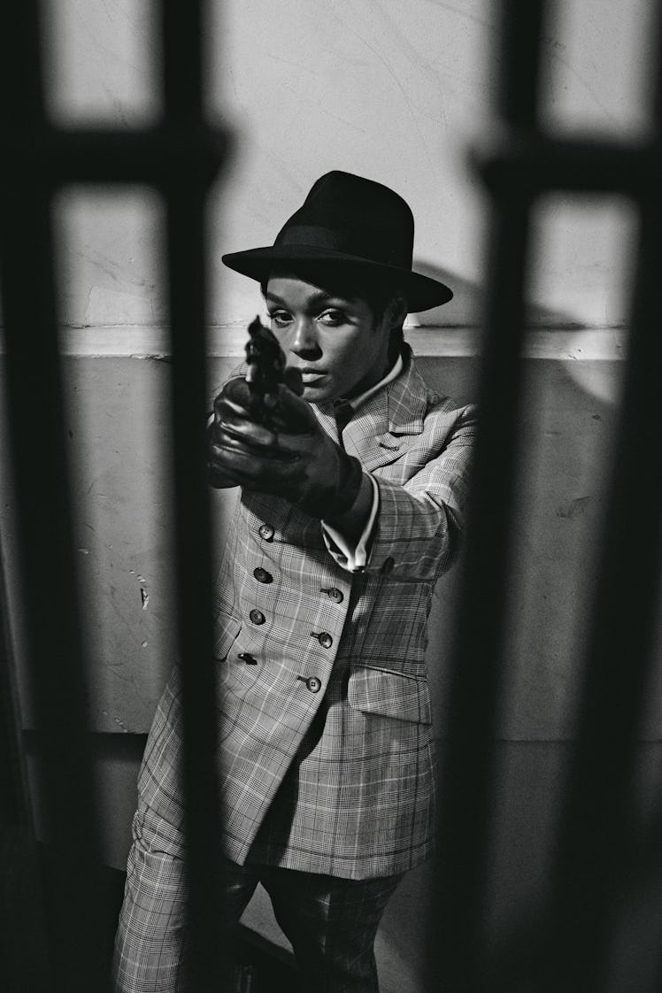 Janelle Monáe posing in a formal suit and a black hat while holding a gun