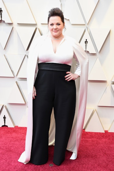 Melissa McCarthy wearing a white cape-top with black pants while posing at the 2019 Oscars Red carpe...