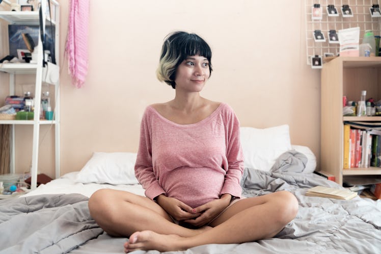 pregnant brown skin asian woman with short highlighted hair sitting on the bed at room smiling posit...