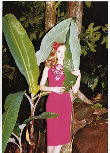 a model in a red dress hiding behind a large tropical plant