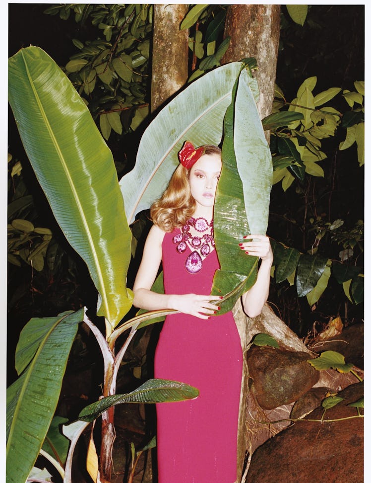 a model in a red dress hiding behind a large tropical plant