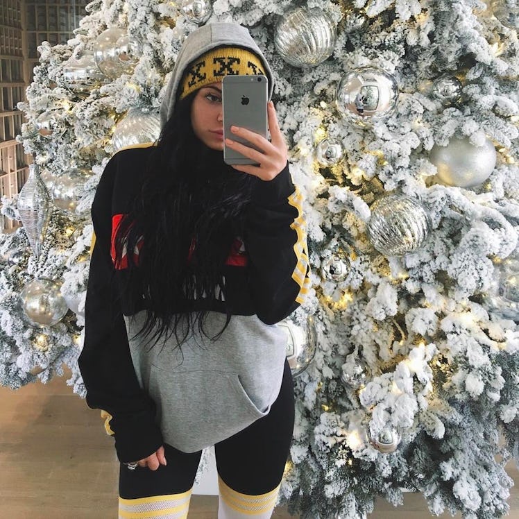 Kylie Jenner taking a mirror selfie in a knit hoodie next to a Christmas tree with white ornaments