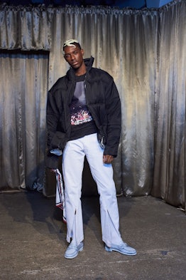 A male model posing while wearing a black jacket and white pants