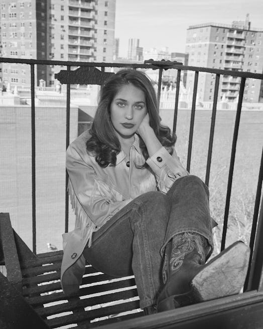 Lola Kirke in a white top, peach jacket and blue denim jeans sitting and posing in black-and-white