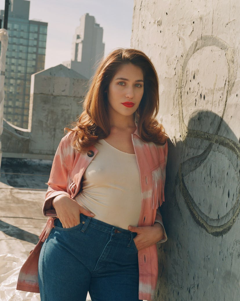 Lola Kirke in a white top, peach jacket and blue denim jeans leaning against a wall