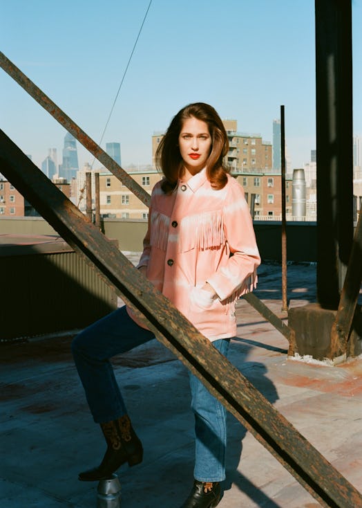 Lola Kirke in a white top, peach jacket and blue denim jeans standing and posing on a rooftop