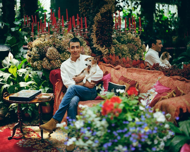 Raqib Shaw holding a dog while sitting on a couch surrounded by flowers.