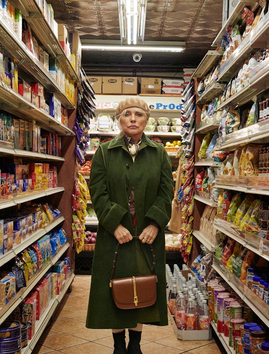 Debbie Harry in a supermarket wearing a green Gucci coat and brown bag with a beige Clyde hat 