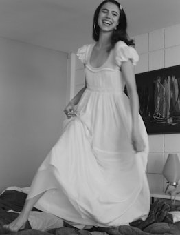 Margaret Qualley laughing while dancing on a bed in a white Chloé dress
