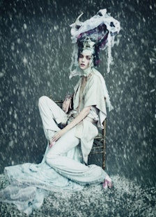 A model posing in a white jumpsuit and sitting on a stool with an abstract headpiece and rain effect