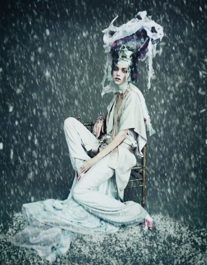 A model posing in a white jumpsuit and sitting on a stool with an abstract headpiece and rain effect