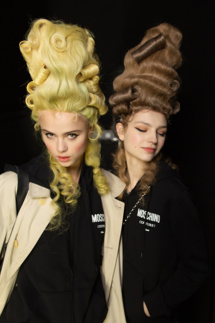 A blonde and a brunette model with high top curly wigs backstage at the Moschino Fall 2020 show