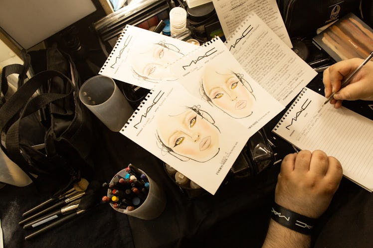 Three makeup sketches on a table with makeup and a person writing something backstage Richard Quinn‘...