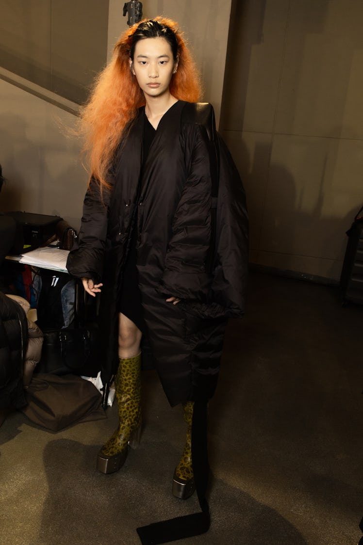 A model in a black asymmetric puffer coat and brown platform boots backstage at the Rick Owens fashi...