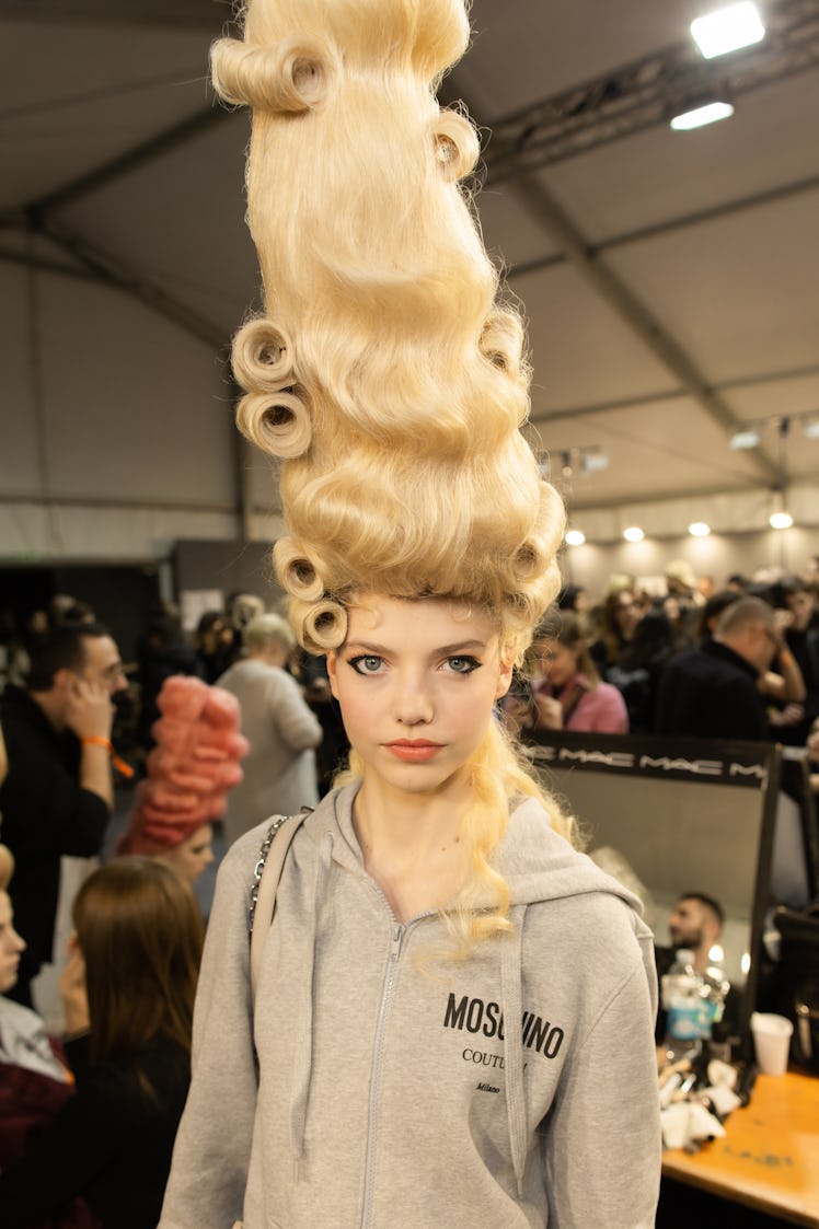 A model with a large curly blonde wig in a grey hoodie backstage at the Moschino Fall 2020 show