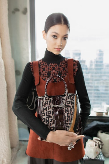 A female model posing while wearing a black shirt and brown dress over it and while holding a brown ...