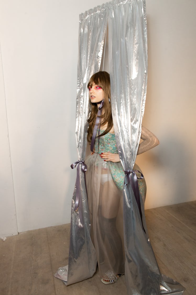 A model in a mint corset and tulle overlay and silver curtains backstage at the Matty Bovan fashion ...