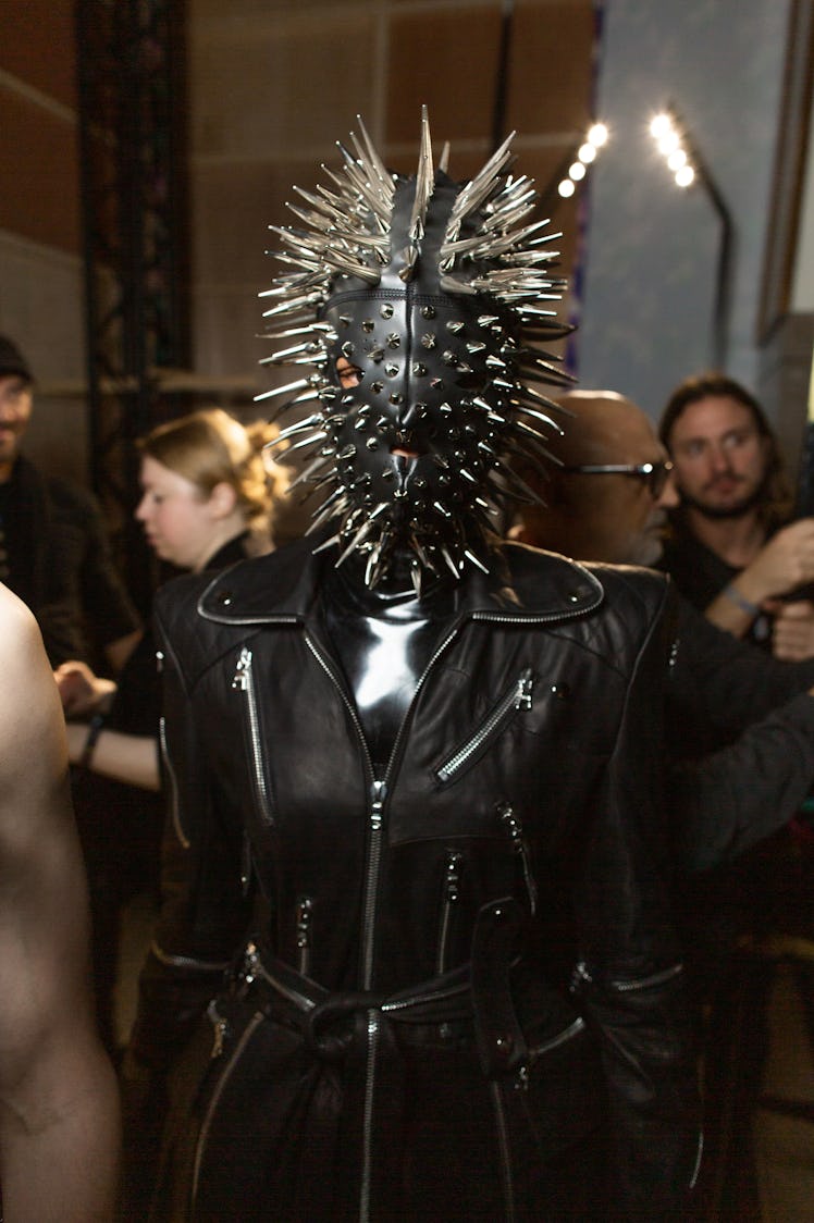 A model in a black jacket, latex top and a latex face mask with spikes backstage at Richard Quinn‘s ...