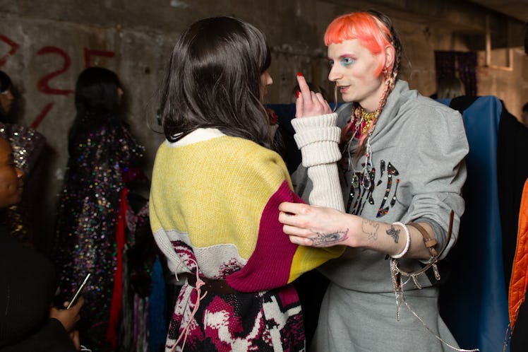 A woman in a yellow-burgundy sweater talking to a person in a grey coat backstage at the Matty Bovan...