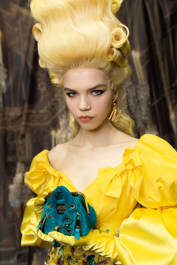 A model with a yellow wig in a yellow frill dress holding a teal bag backstage at the Moschino Fall ...