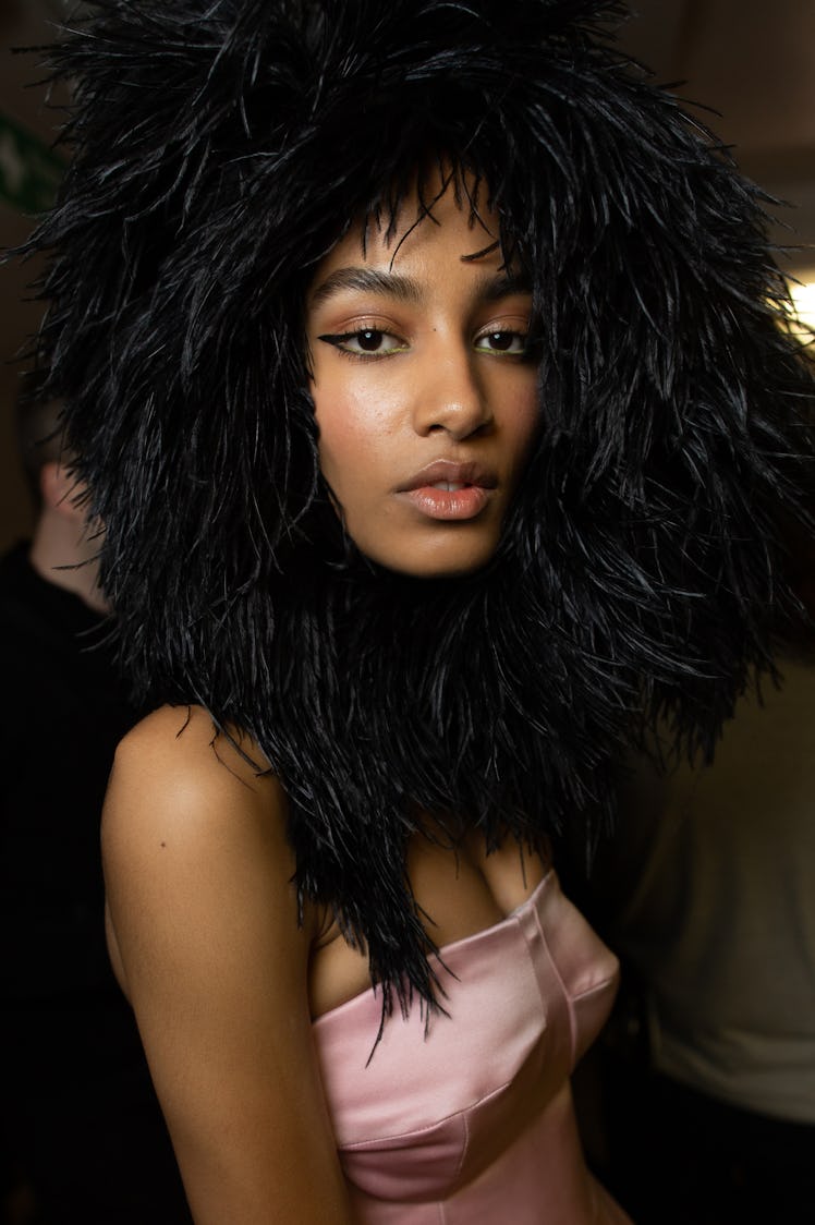 A close-up portrait of a model in a pink satin top and a black feather hat backstage at Richard Quin...