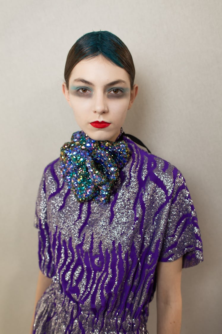 A female model posing in a sequined purple and silver dress while having white makeup on her face