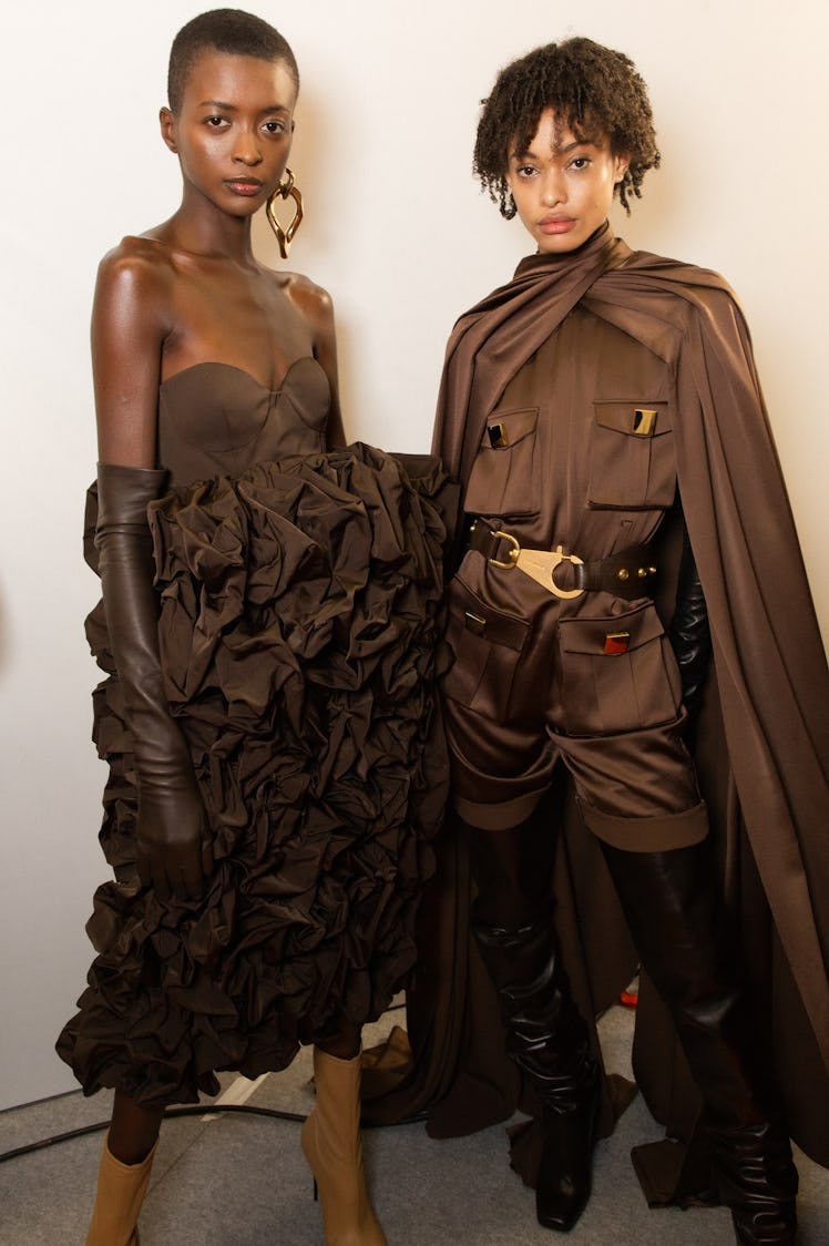 A model in a black puff dress and a model in a brown coat backstage at Balmain Fall 2020