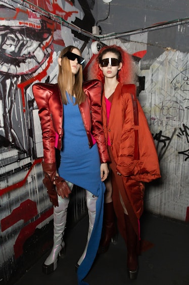 A model in a blue dress and red jacket and a model in a red coat backstage at the Rick Owens fashion...