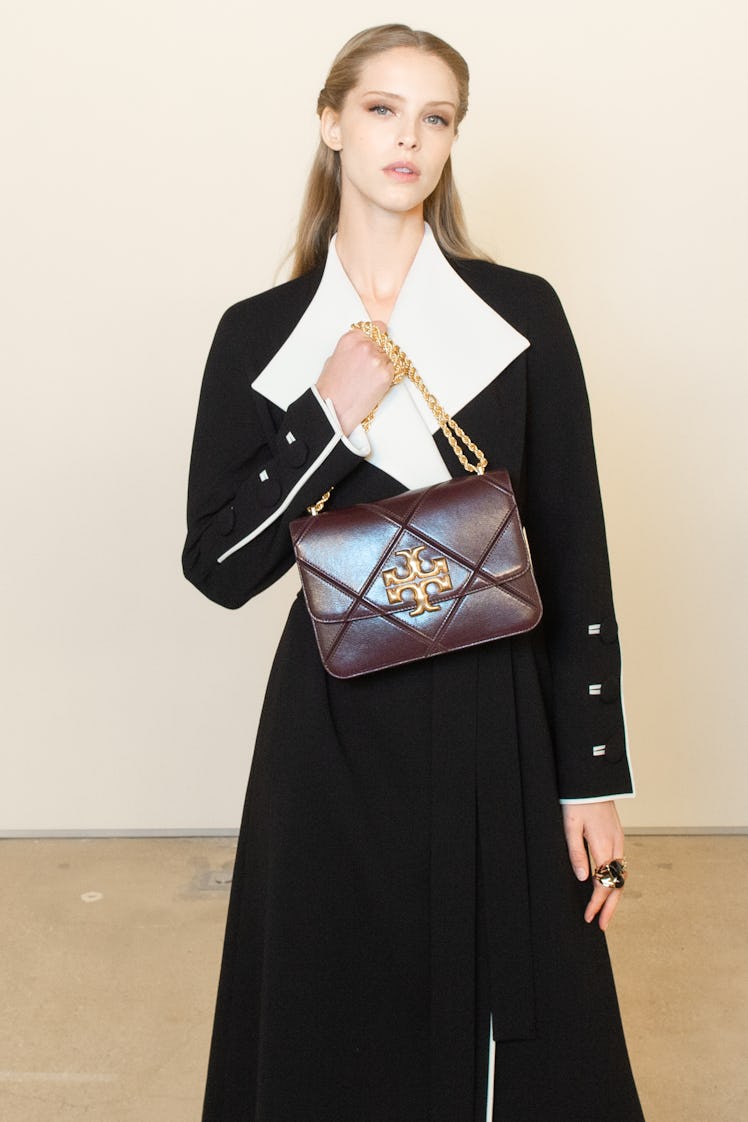 A blonde model posing while wearing a black coat and while holding a burgundy bag